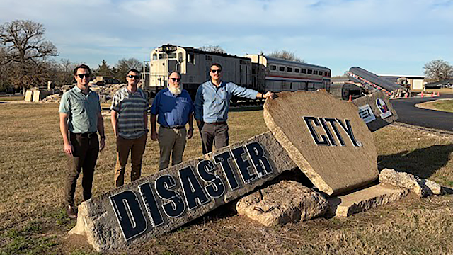 Four men stand outdoors in front of a large broken stone sign that reads "Disaster City," with a staged train wreckage visible in the background.